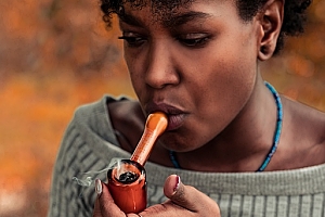 Black woman with grey boat neck sweater smokes out of red cannabis glass bowl